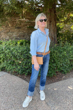 blonde woman weaing chambrays shirt, cuffed denim and golde sneakers standing in front of tree