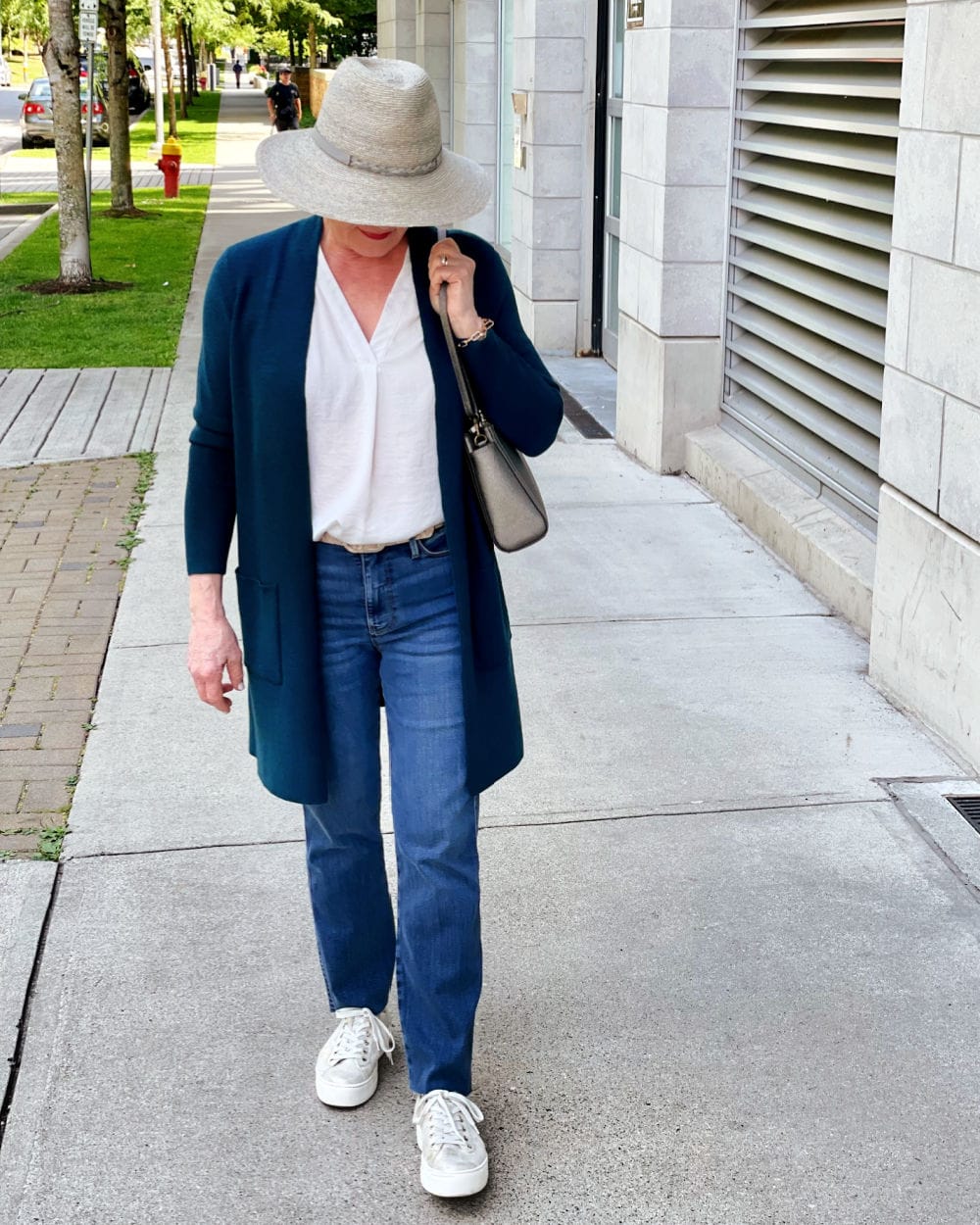 woman wearing hat and long sweater with casual denim walking down sidewalk