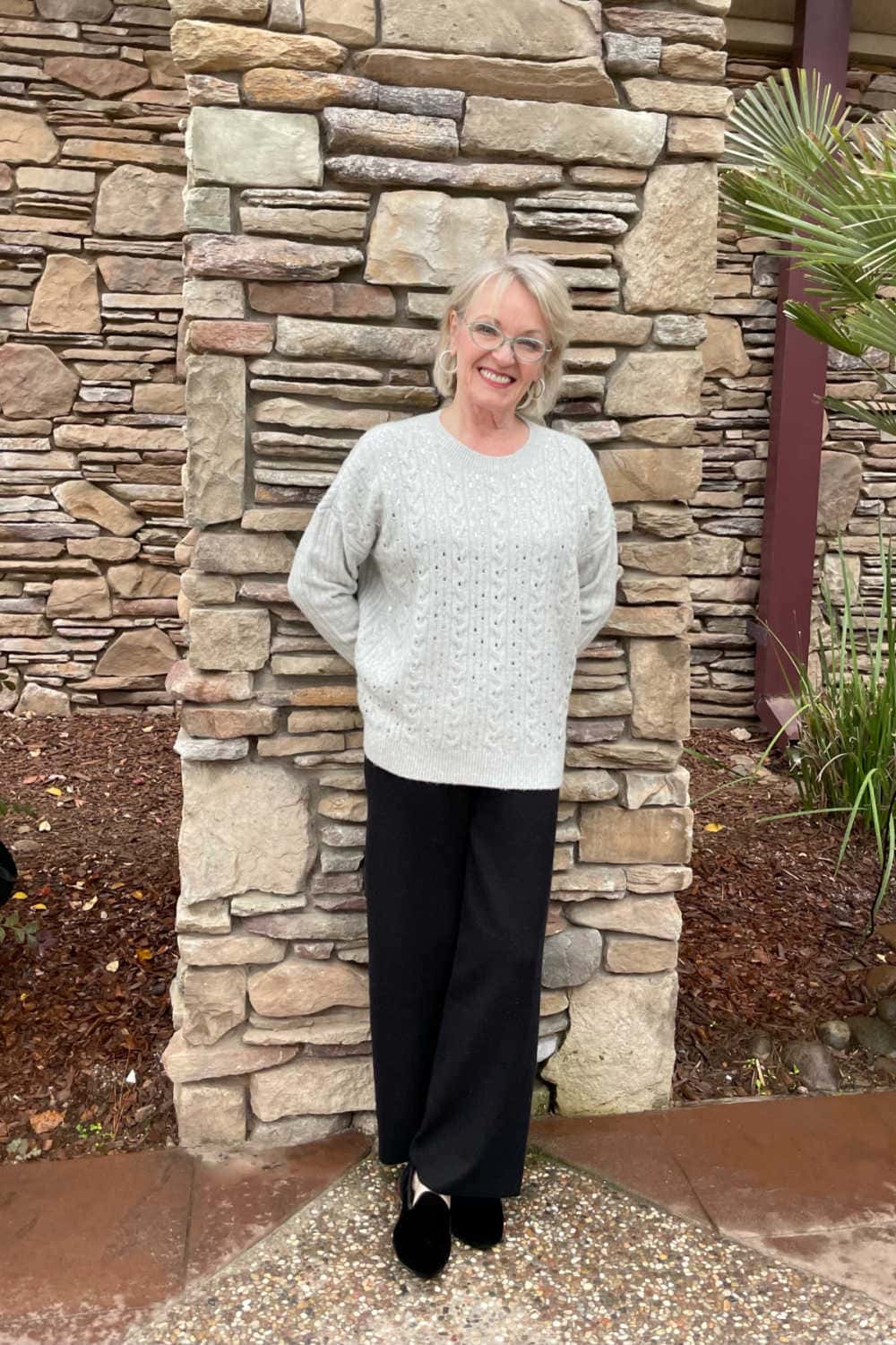 woman standing againt wall wearing gray sweater and black pants