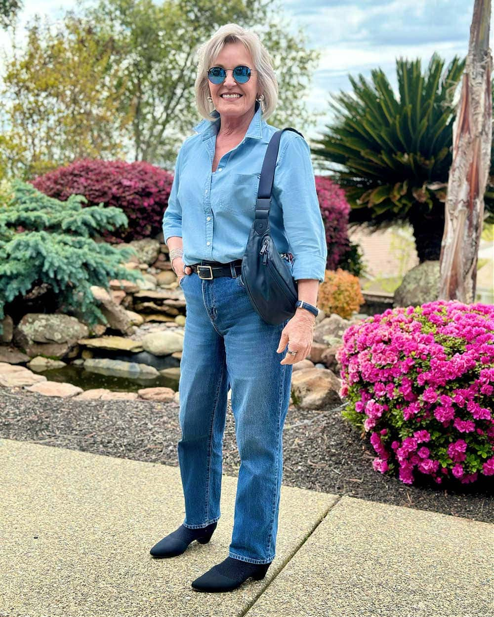 Jennifer wearing a light blue denim shirt tucked into dark wash jeans with a navy crossbody bag and sunglasses.