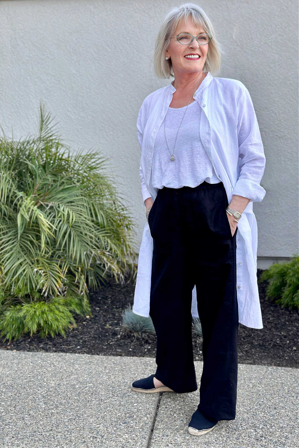 women over 60 wearing black linen pants, white linen tank and white linen duster with simple pearl earrings