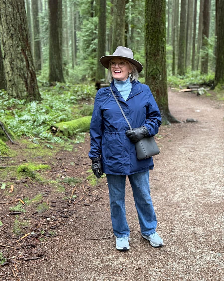 Jennifer Connolly wearing a blue Lands’ End Squall Jacket with jeans, a gray hat, gloves, and a crossbody bag while walking on a wooded trail.