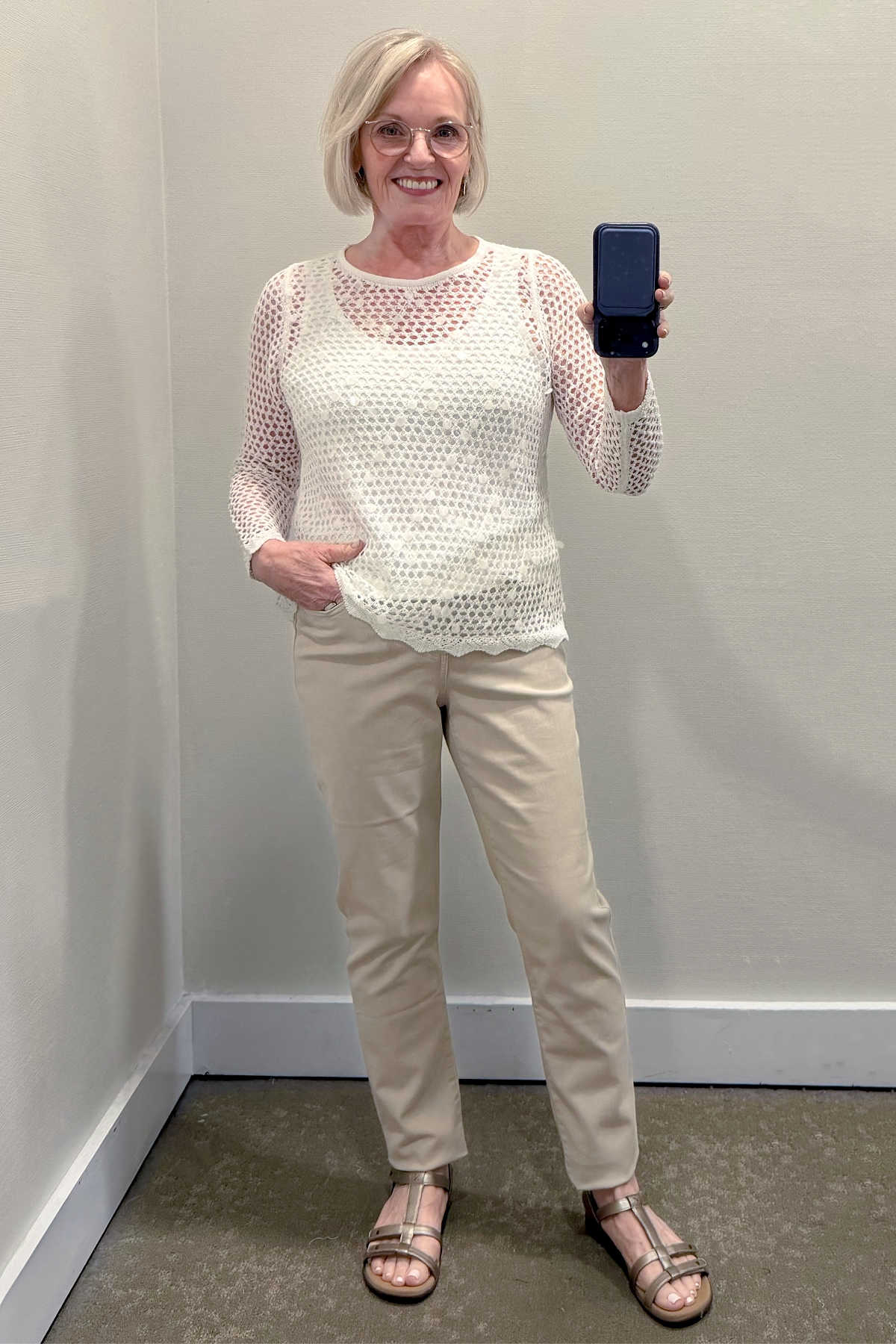 Jennifer Connolly wearing a white open-knit sweater over a camisole with cream ankle pants and metallic sandals for summer.